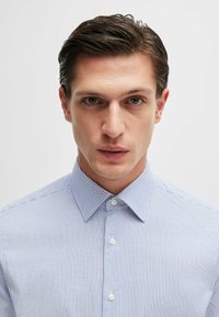 Camicia a righe azzurro chiaro e bianco con colletto, abbottonata sul davanti e bottoni bianchi semplici. Il tessuto appare liscio e ben rifinito.