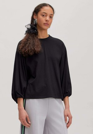 Femme aux cheveux bouclés attachés avec un chouchou noir, portant de grandes boucles d’oreilles anneaux, un haut noir à manches longues et un pantalon gris clair avec des bandes latérales.