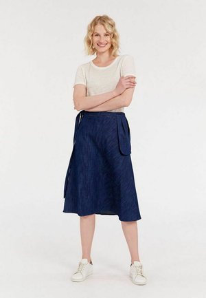 T-shirt ajusté gris clair associé à une jupe portefeuille bleu foncé avec un ourlet asymétrique et une poche latérale, portée avec des baskets blanches.