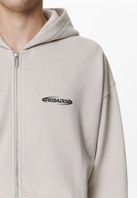 Sudadera con capucha color beige claro con cremallera, con un logo negro de "PEGADOR" en el pecho y tela suave y texturizada.
