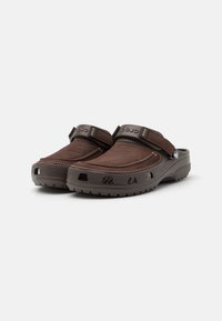Mules bruns à enfiler avec un dessus doux, sangle ajustable, surface texturée et trous de ventilation sur les côtés. Semelle en caoutchouc épaisse.
