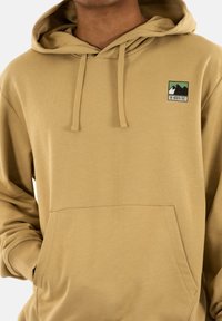 Sudadera con capucha color marrón claro, con un bolsillo frontal, cordones y un parche de logo con montañas. Fabricada con un tejido suave y texturizado.