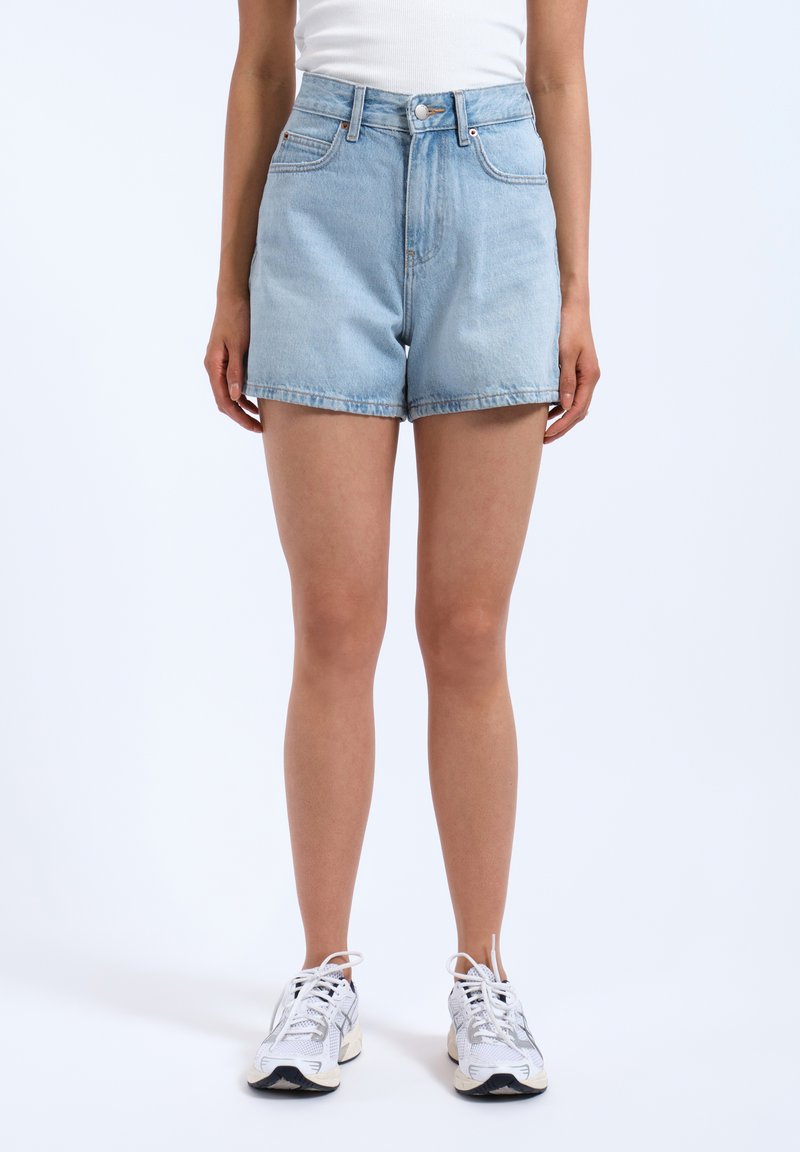 Hellblaue Jeansshorts mit hoher Taille, ausgestattet mit Fronttaschen, einem Knopfverschluss und einer lässigen Passform. Kombiniert mit weißen Turnschuhen.