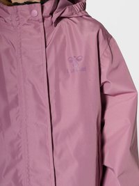 Nahaufnahme einer Person, die eine hellviolette wasserdichte Jacke mit Kapuze und kleinem "hummel"-Logo auf der Brust trägt.
