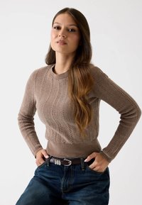 Brauner Kabelstrickpullover mit langen Ärmeln, gerippten Bündchen und taillierter Passform, kombiniert mit hochtaillierten blauen Jeans und einem braunen Gürtel.