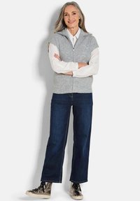 Graue ärmellose Zip-Weste über einem weißen Langarmshirt, kombiniert mit dunkelblauen Weitbein-Jeans und metallischen Sneakers.