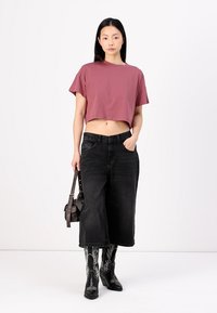 Camiseta maroon curta, de mangas curtas, combinada com culottes largas pretas e botas de cowboy pretas com padrão. A segurar uma pequena mala castanha.