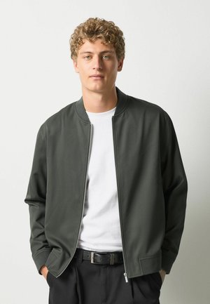 Ung mand med krøllet blond hår iført en mørkegrøn jakke med lynlås, hvid t-shirt og sorte bukser med hænderne i lommerne.