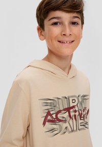 Béžová mikina s kapucí a předním grafickým potiskem v tmavě červené a černé barvě. Měkká látka, uvolněný střih a kapuce. Významné detaily zahrnují tučné písmo.