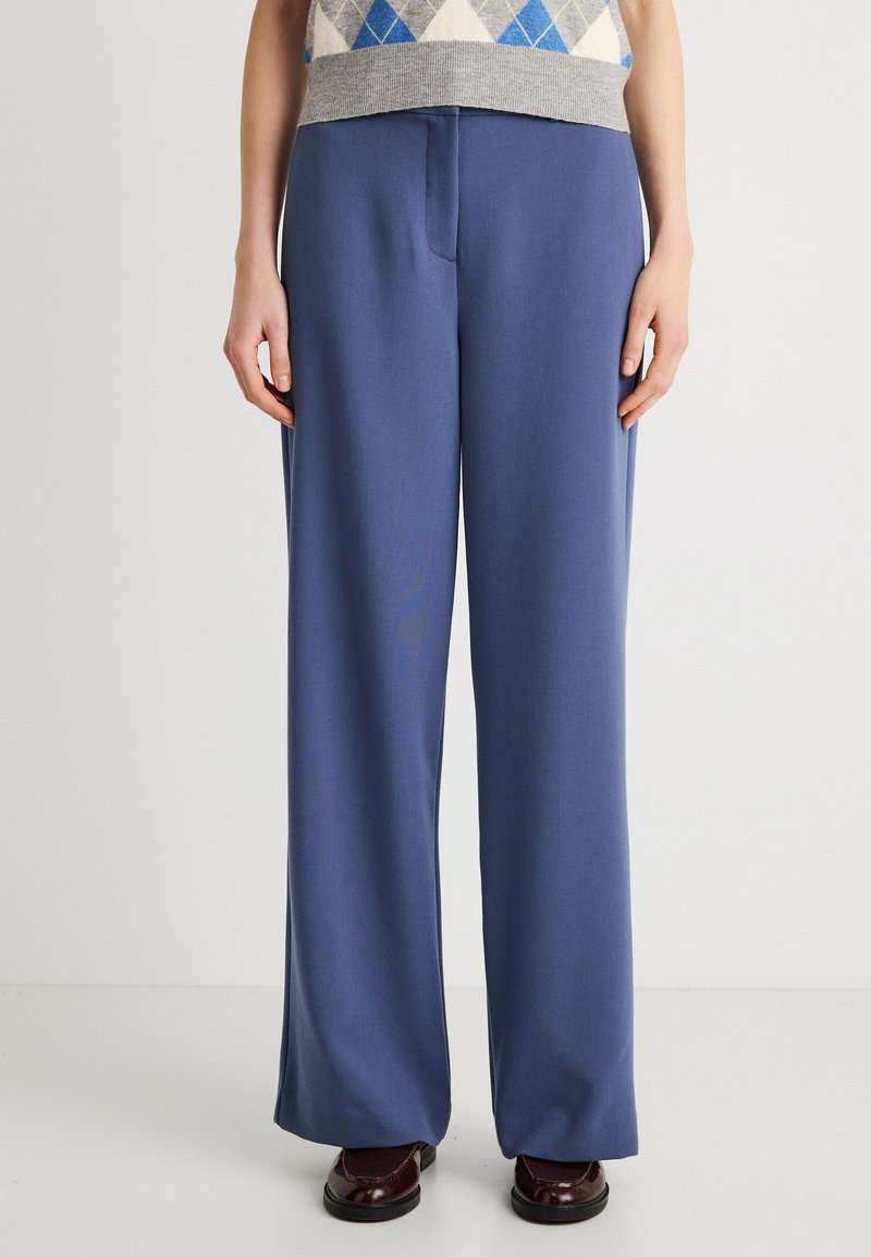 Παντελόνια με φαρδιά πόδια, κατασκευασμένα από λεία, μπλε ύφασμα, με πιέτα μπροστά και κλασική γραμμή. Συνδυασμένα με σκούρα παπούτσια.