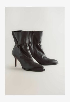 Bottines noires en cuir verni à talons hauts avec des pointes pointues et des fermetures éclair sur le côté, présentées contre un fond clair et uni.