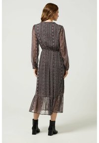 Largo vestido con estampado que tiene una base oscura, con diseños florales en rosa, cintura ceñida, mangas largas y un dobladillo con volantes. Combinado con botas negras hasta el tobillo.