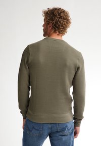 Felpa verde oliva con tessuto testurizzato, polsini e vita a costine, scollo rotondo e vestibilità casual, abbinata a jeans blu.