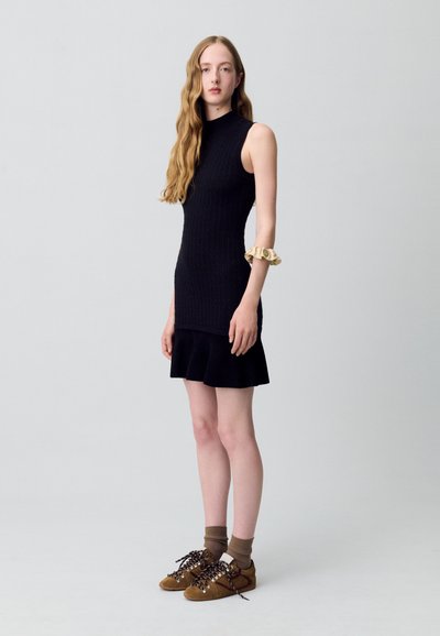 Abito nero senza maniche lavorato a maglia con corpetto aderente e orlo svasato, abbinato a scarpe in suede marrone e calze marroni. Scrunchie dorato al braccio.
