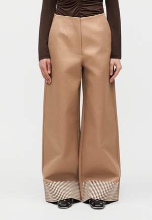 Φαρδιά καφέ παντελόνια με καρό μανσέτες, φορεμένα με μαύρα παπούτσια και καφέ μακρυμάνικο τοπ με ρυτιδωτή λεπτομέρεια, εμπρόσθια όψη σε ουδέτερο φόντο.