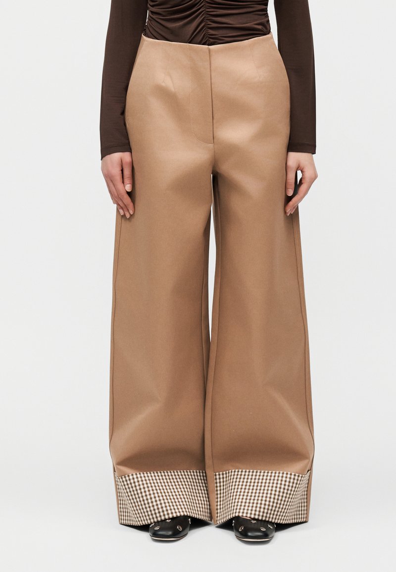 Φαρδιά καφέ παντελόνια με καρό μανσέτες, φορεμένα με μαύρα παπούτσια και καφέ μακρυμάνικο τοπ με ρυτιδωτή λεπτομέρεια, εμπρόσθια όψη σε ουδέτερο φόντο.