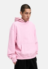 Helle rosa Kapuzenjacke aus weichem Baumwollmaterial. Verfügt über eine frontale Tasche, eine Kordelzugkapuze und gerippte Manschetten. Kombiniert mit schwarzen Hosen.