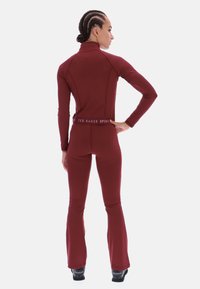 Tumši sarkana sporta apģērba komplekts ar garām piedurknēm un izplestām biksēm; izgatavots no elastīga auduma; ar augstu apkakli un jostasvietas logo.