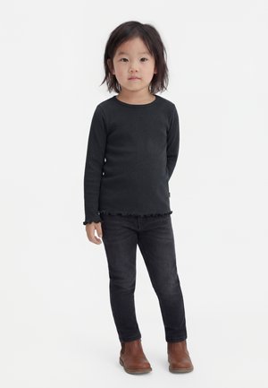 Malé dieťa stojí, má na sebe čierne tričko s dlhým rukávom, čierne džínsy a hnedé členkové topánky na jednoduchom bielom pozadí.