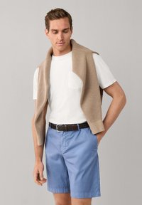 Hombre con suéter beige sobre los hombros, vistiendo camiseta blanca metida en pantalones cortos azules con un cinturón oscuro, de pie contra un fondo gris.