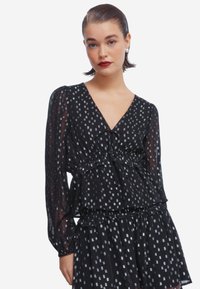 Blusa nera a strati con maniche lunghe trasparenti, caratterizzata da un profondo scollo a V e un motivo geometrico argentato su tutta la superficie. Dettagli arricciati in vita.