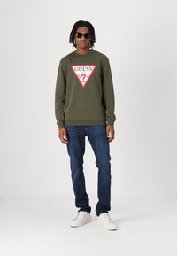 Grønn hettegenser med rødt trekantet logo, paret med blå jeans og hvite joggesko. Modellen har på seg svarte solbriller og står mot en ensfarget bakgrunn.