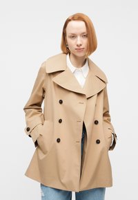 Γυναίκα με κοντά κόκκινα μαλλιά, ντυμένη με μπεζ διπλόμπουτο καμπαρντίνα πάνω από λευκό πουκάμισο και ανοιχτόχρωμα τζιν, στέκεται με τα χέρια στις τσέπες.