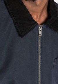 Marinblå jacka med en svart corduroy-krage, metallisk dragkedja och strukturerad design. Texturerad tyg och minimalistisk detaljer.