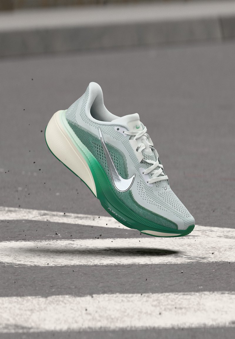 Chaussure de course gris clair et verte avec des lacets blancs et un logo Nike argenté flottant au-dessus d'une route aux bandes blanches.