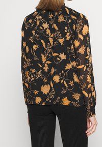 Blouse noire avec un motif floral orange, manches longues et texture lisse. Présente un pli au dos et des poignets élastiques.