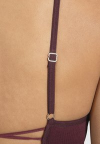 Tirante de sujetador burdeos con un ajustador de metal plateado, que presenta una tela texturizada y un diseño de tirantes cruzados sobre un fondo color piel.