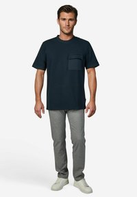 Donkerblauw T-shirt met korte mouwen en een borstzakje, gecombineerd met lichtgrijze slim-fit broek en witte sneakers. Eenvoudig en functioneel ontwerp.