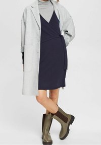 Grå oversized kappa över en marinblå omslagsklänning. Modellen bär gröna gummistövlar med bruna elastiska detaljer. Slät tygtextur och lagrad design.