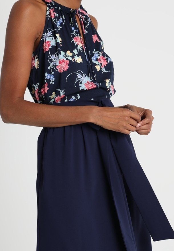 Femme portant une robe sans manches bleu marine avec un haut à col halter fleuri et une ceinture nouée à la taille, les mains reposant près de la taille.