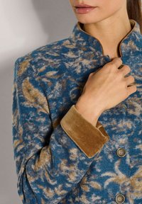 Vrouw in een blauw en tan patroon jas met fluwelen manchet en mandaringkleur, die met haar hand de bovenste knoop vastmaakt.