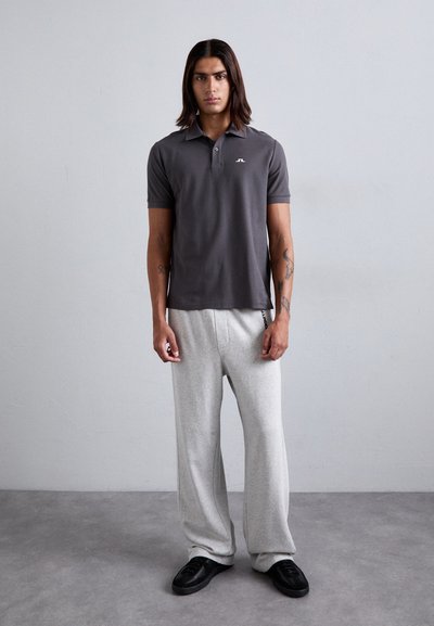 Graues Polohemd mit einem dezenten Logo, kombiniert mit hellgrauen, locker sitzenden Jogginghosen und schwarzen Schuhen, vor einem neutralen Hintergrund stehend.
