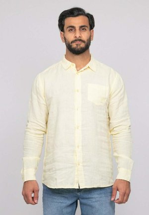 Homme aux cheveux foncés et à la barbe, portant une chemise jaune pâle à manches longues boutonnée et un jean bleu, debout devant un fond clair uni.