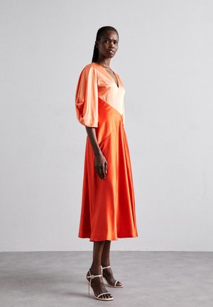 Orange midi-klänning med v-ringning, puffärmar och flödande silhuett; bärs med remmar och prydda högklackade sandaler.