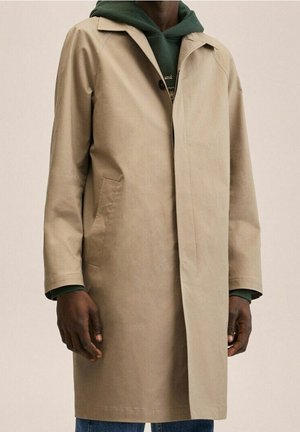 Homme portant un manteau beige mi-long sur un sweat à capuche vert et un jean bleu sur fond uni.