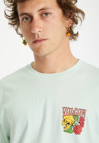 Camiseta de manga corta verde menta con un gráfico de una calavera amarilla y flores en el pecho izquierdo. Tela suave con un cuello redondo y un ajuste relajado.