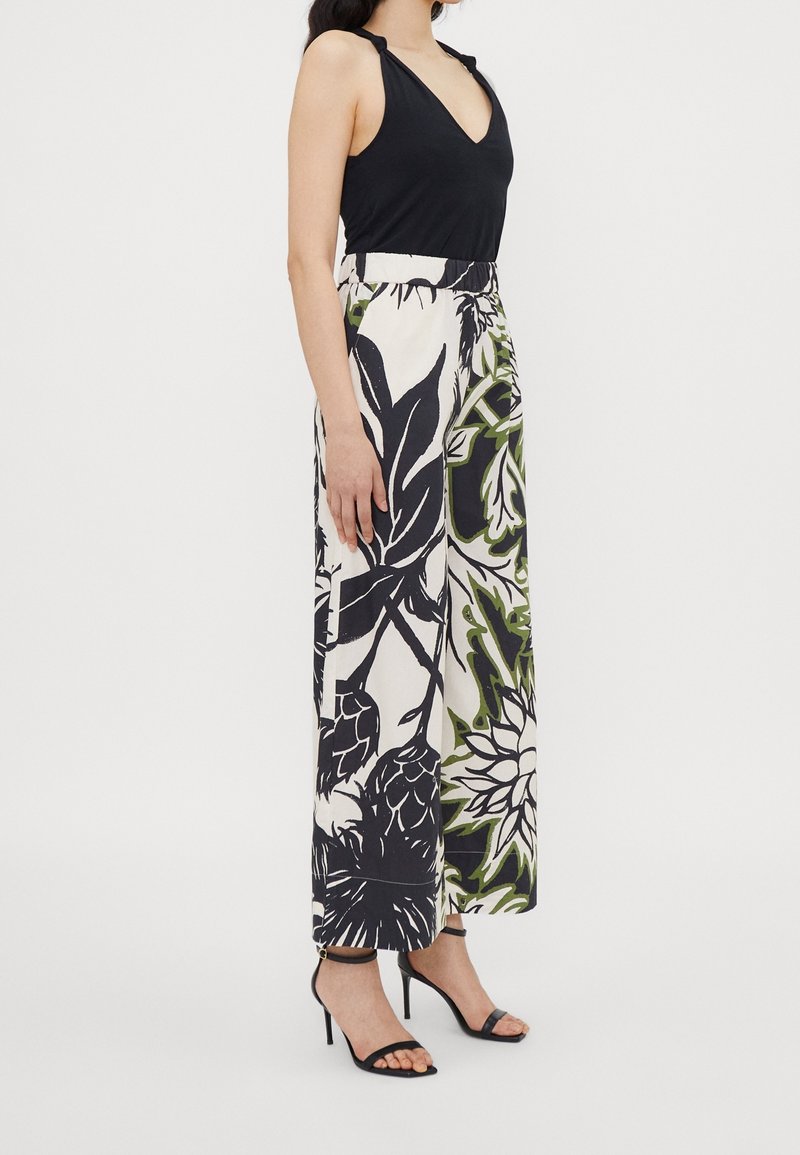 Femme portant un haut noir sans manches et un pantalon blanc à jambes larges avec un grand imprimé floral noir et vert, associé à des talons noirs à brides.