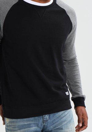 Homme portant un sweat-shirt raglan noir et gris associé à un jean bleu clair déchiré sur fond uni.