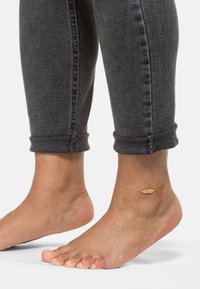 Gul ankelkæde med en muslingekonformet vedhæng, båret på en bar ankel. Grå denim, rullet ved kanten, med synlige syningsdetaljer.