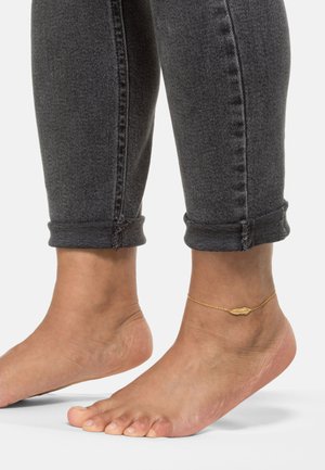 Goldene Fußkette mit einem muschelförmigen Anhänger, getragen an einem nackten Knöchel. Graue Jeans mit umgeschlagenem Saum und sichtbaren Nähdetails.