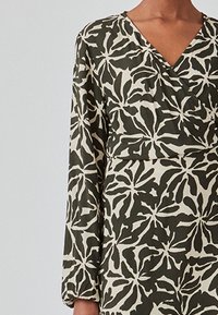 Mønstret kjole med et mørkegrønt og cremefarvet bladdesign, v-hals, lange ærmer og en tætsiddende talje. Stoffet har en glat tekstur.
