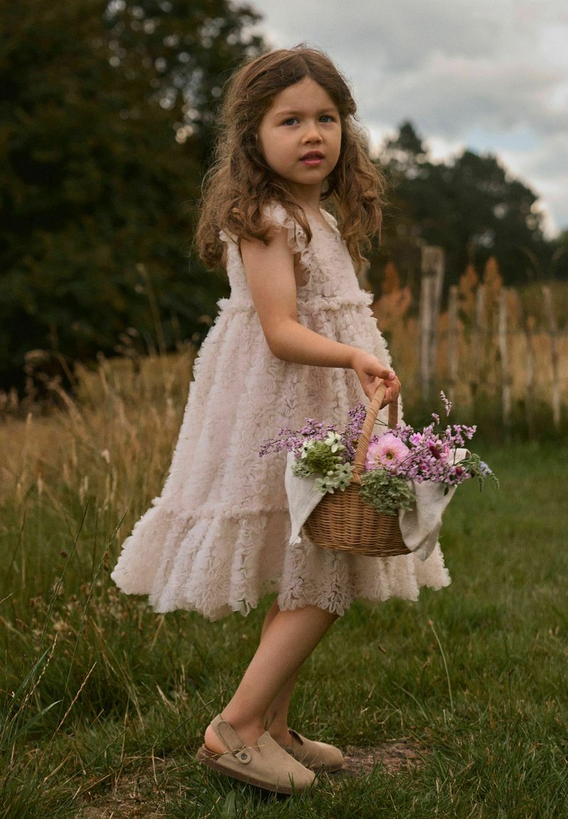Jeune fille en robe pâle à volants tenant un panier en osier rempli de fleurs roses et violettes, debout dans un champ herbeux avec des arbres et un ciel nuageux.