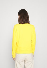 Sudadera amarilla brillante con mangas largas, puños acanalados y cuello redondo. Textura de tela suave, combinada con pantalones de color claro.
