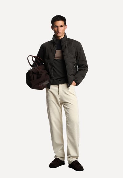 Giovane uomo con giacca a vento nera, maglione a righe, pantaloni di corduroy crema, mocassini scuri, che tiene una grande borsa di pelle e tessuto marrone.