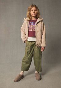 Jachetă din fleece bej cu glugă, tricou cu imprimeu bordo, pantaloni cargo verde măsliniu, șosete bej și încălțăminte maro slip-on.