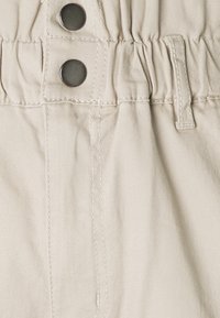 Gros plan sur la ceinture d'un pantalon en tissu beige avec deux boutons-pression métalliques, fronces élastiques, passants de ceinture et détails de couture visibles.